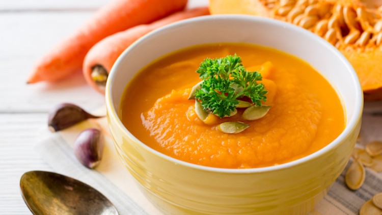 Eine gelbe Schale mit orangefarbener Kürbissuppe, dekoriert mit Kürbiskernen und Petersilie, daneben Karotten und Knoblauch.