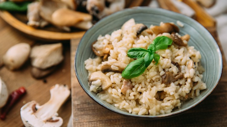 Ein Teller cremiges Pilzrisotto mit frischen Champignons und Basilikum-Zweig, umgeben von rohen Pilzen auf Holzuntergrund.