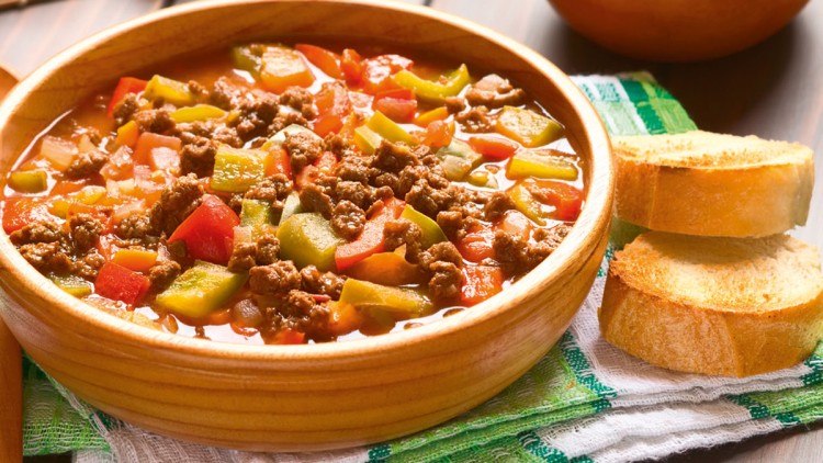 Ein Teller mit Suppe aus Hackfleisch, Paprikastücken und Tomaten, daneben zwei Scheiben geröstetes Weißbrot auf einem Stofftuch.