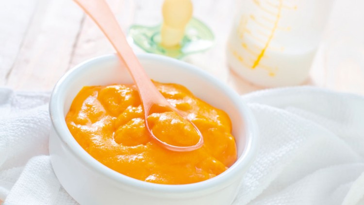 Weiße Schale mit orangefarbenem Brei und transparentem Kunststofflöffel, daneben Schnuller und Babyflasche im Hintergrund.