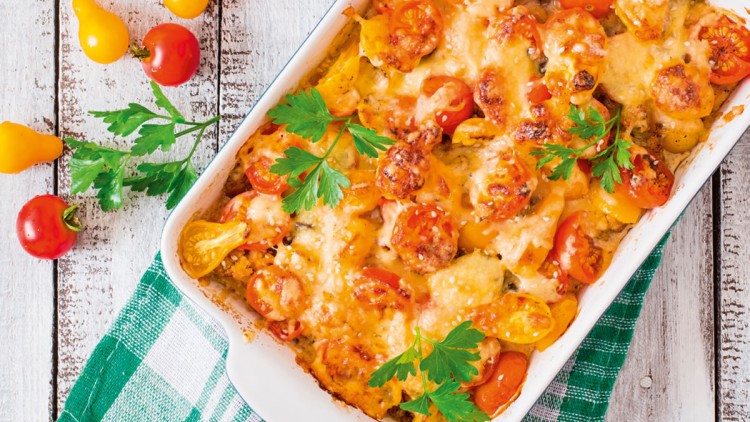 Auflauf mit geschmolzenem Käse, gelben und roten Tomaten sowie Petersilie in einer weißen Auflaufform auf Holztisch.
