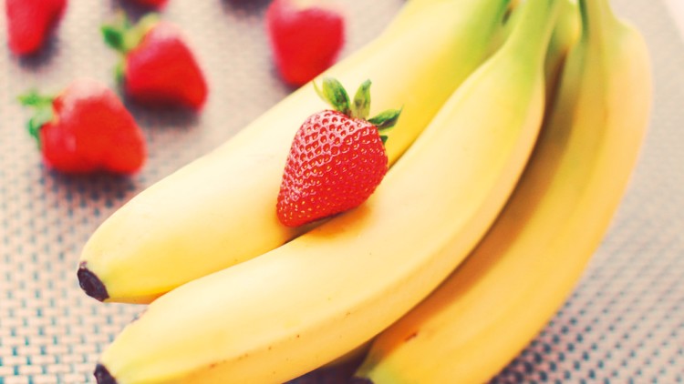 Drei gelbe Bananen liegen zusammen, vorne liegt eine rote Erdbeere, im Hintergrund sind weitere unscharfe Erdbeeren zu sehen.