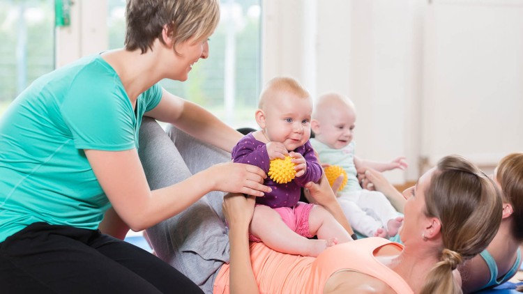 Erwachsene und Babys sitzen oder liegen auf bunten Matten, ein Baby hält einen gelben Massageball in den Händen.
