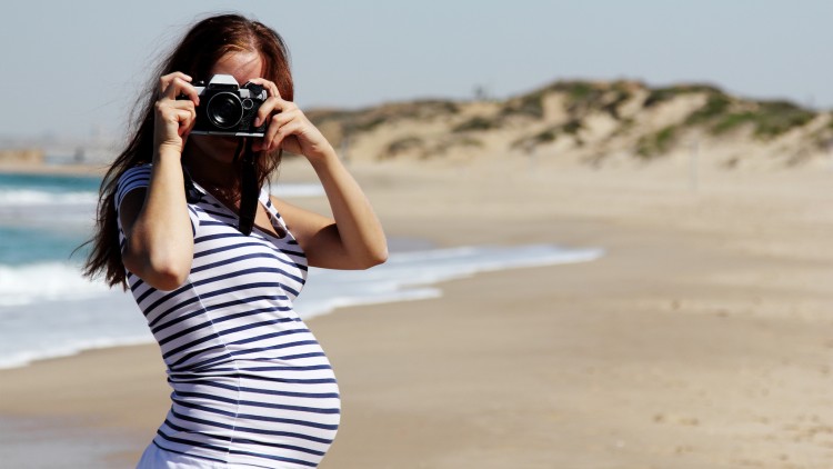 Person in gestreiftem Oberteil und weißer Hose steht am Sandstrand, dahinter das Meer und Dünen im Hintergrund.