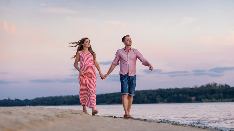 Zwei Personen halten sich an den Händen und spazieren am Sandstrand entlang, im Hintergrund Wasser und ein Himmel bei Sonnenuntergang.