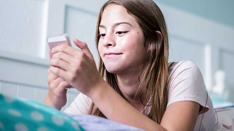 Person liegt auf einem Bett, trägt Jeans und ein helles Oberteil und hält ein Smartphone in beiden Händen.