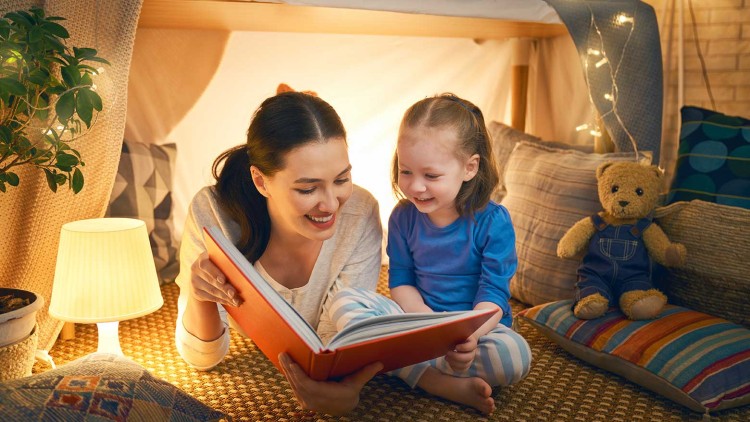 Eine Person liest mit einem Kind aus einem orangefarbenen Buch vor, umgeben von Kissen, Decken und einer Lampe.
