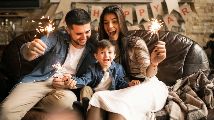 Vier Personen sitzen auf einem Ledersofa vor einer Winterdeko und halten Wunderkerzen, im Hintergrund steht „HAPPY NEW YEAR“.