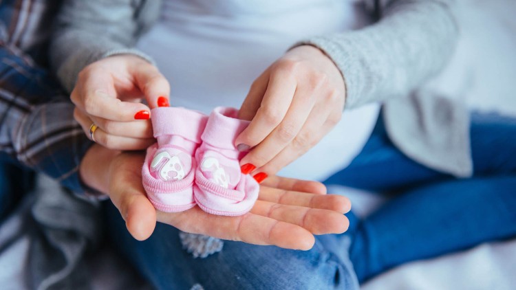 Zwei Hände halten rosa Babyschuhe mit aufgedrucktem Hundemotiv, die auf einer geöffneten Handfläche liegen.