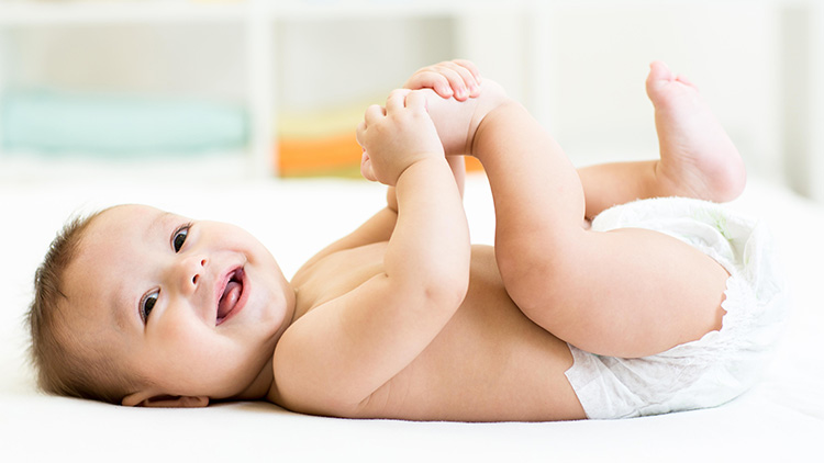 Ein Baby liegt auf dem Rücken auf einer weißen Unterlage und hält mit beiden Händen seine Füße, es trägt eine Windel.