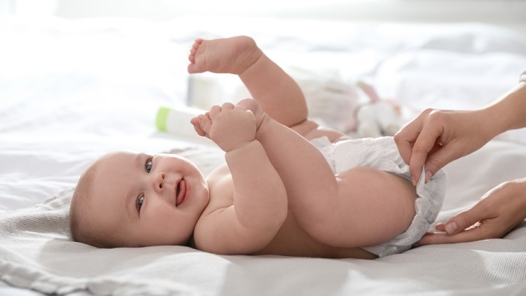 Ein Baby liegt auf dem Rücken, eine Hand wechselt die Windel, im Hintergrund sind Babyutensilien erkennbar.