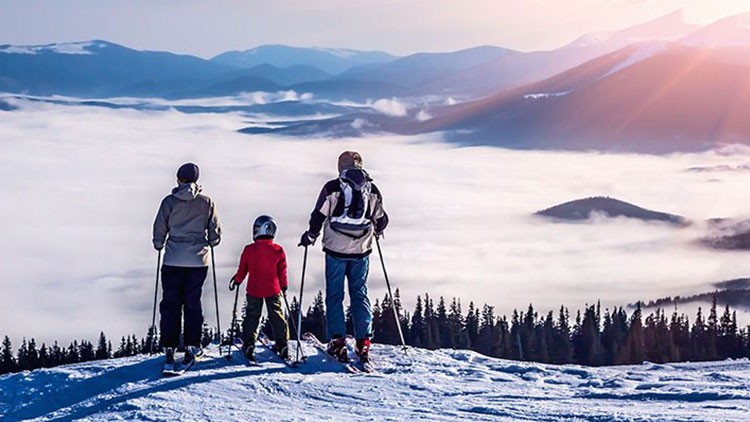 Drei Personen in Wintersportkleidung stehen mit Skiern auf einem schneebedeckten Hang und blicken auf eine nebelige Berglandschaft.