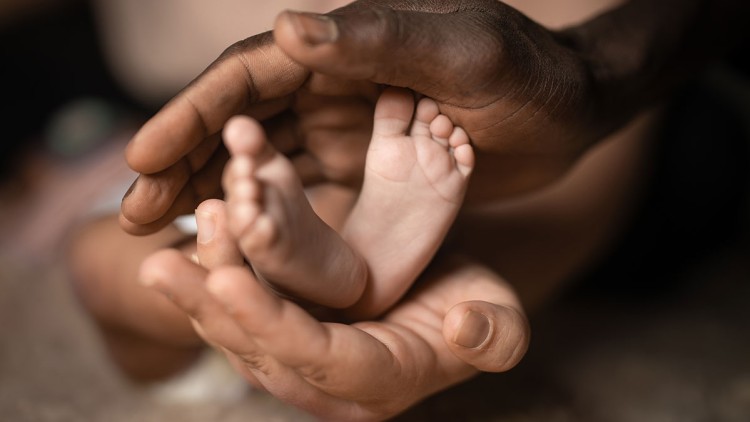 Zwei erwachsene Hände halten behutsam die kleinen Füße eines Babys, weicher Untergrund im Hintergrund.