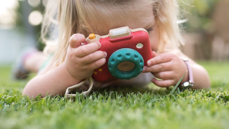 Kind liegt auf Gras und hält eine rot-grüne Spielzeugkamera mit gelbem Knopf und blauem Objektiv vor das Gesicht.
