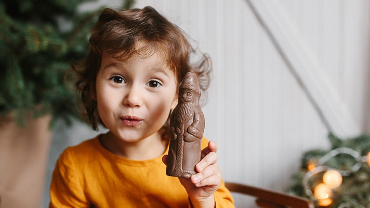 Eine Person in orangefarbener Kleidung hält einen Schokoladen-Weihnachtsmann, im Hintergrund sind Tannengrün und Lichterketten.