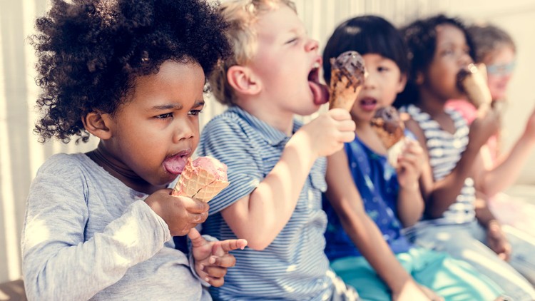 Kinder sitzen nebeneinander und essen Eiswaffeln, Kleidung in Blau- und Grautönen, Hintergrund mit Sonnenlicht.