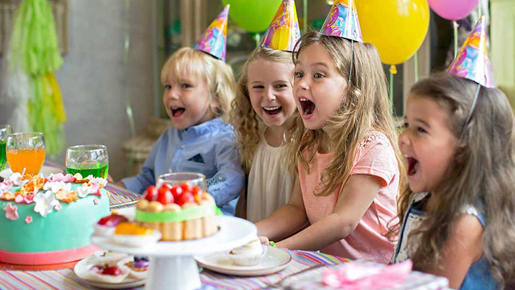 Kinder sitzen an einem Tisch mit Getränken, einem Kuchen mit Blumen-Dekoration und Luftballons im Hintergrund.