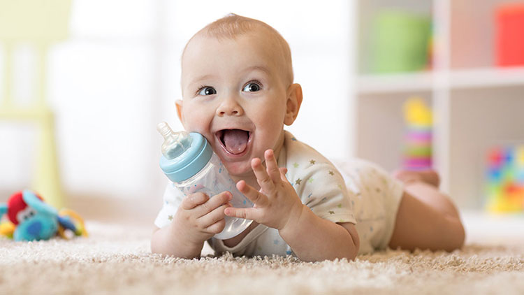 Ein Baby in hellem Strampler liegt auf dem Bauch und hält eine Babyflasche, im Hintergrund unscharfes Spielzeug und Möbel.
