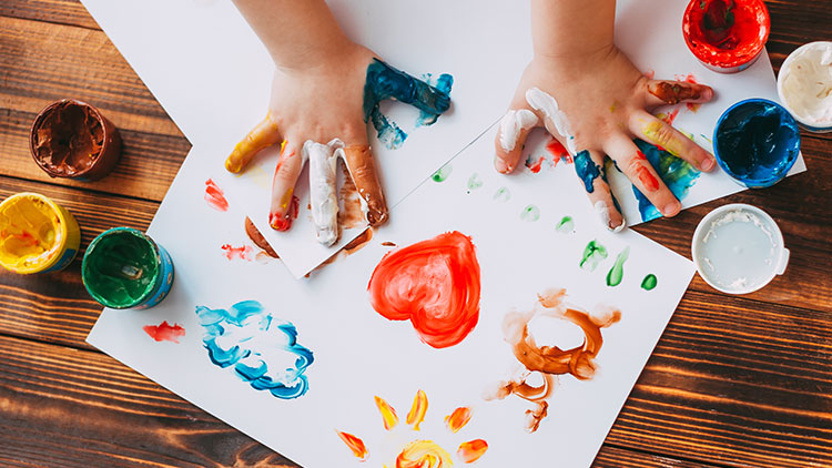 Kinderhände malen mit Fingerfarben auf weißem Papier, bunte Farbtöpfe stehen auf einer Holzoberfläche.