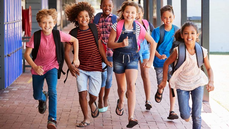 Eine Gruppe von Kindern mit Rucksäcken rennt lachend durch einen überdachten Schulflur aus Backstein.