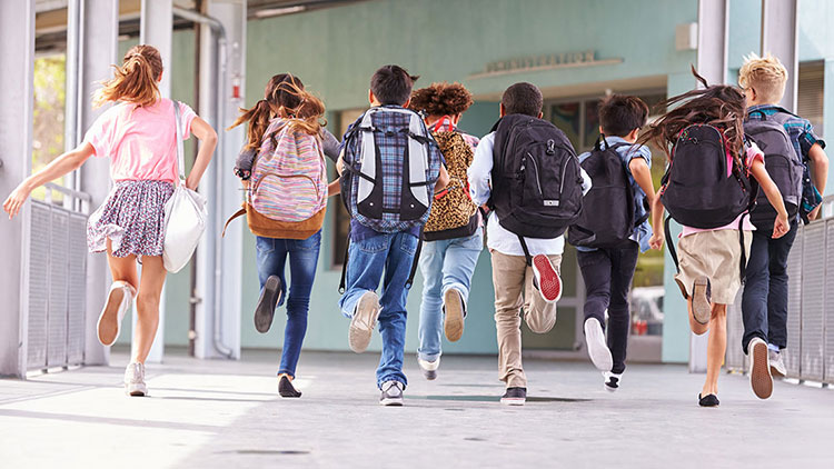 Eine Gruppe von zehn Kindern mit Rucksäcken läuft einen überdachten Weg entlang auf ein Gebäude mit Glasfenstern zu.