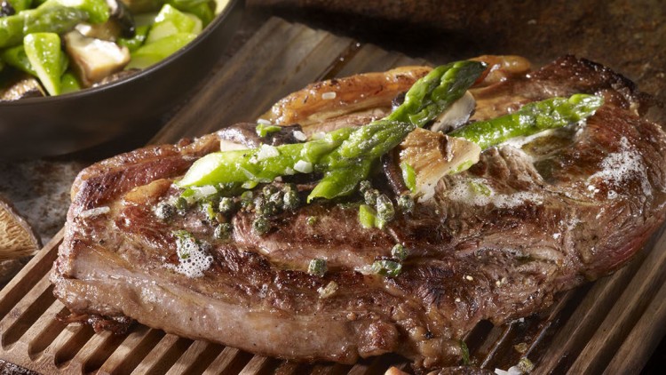 Gegrilltes Steak mit grünem Spargel und Pilzen auf Holzunterlage, daneben eine Schüssel mit Gemüse und frischen Pilzen.