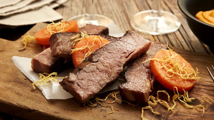Gebratene Fleischscheiben und Karottenscheiben mit knusprigen Garnituren auf Holzbrett, daneben Besteck und Schälchen mit Püree.