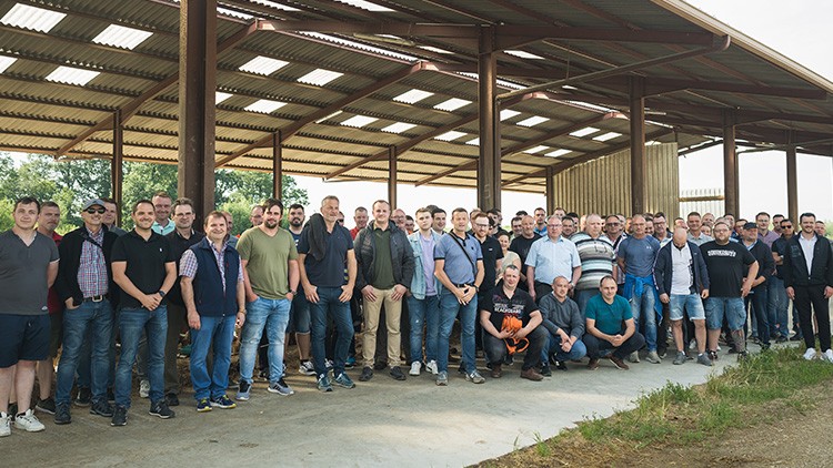 Gruppe von Menschen steht in einer offenen Halle mit Wellblechdach und Pfeilern, im Hintergrund Bäume und Himmel sichtbar.