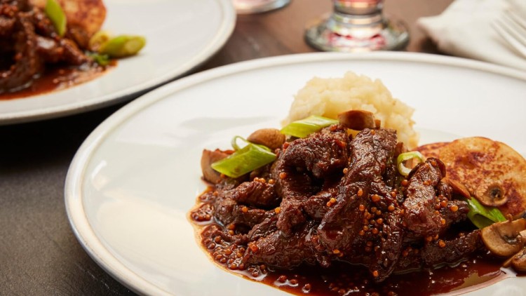 Ein Teller mit Rindfleisch in dunkler Sauce, Pilzen, Frühlingszwiebeln, Kartoffelpürree und einem Kartoffelpuffer.