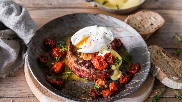 Ein gegrilltes Rindfleisch-Patty mit pochiertem Ei, getrockneten Tomaten und Sauce auf einem Teller, daneben Brot und ein Tuch.