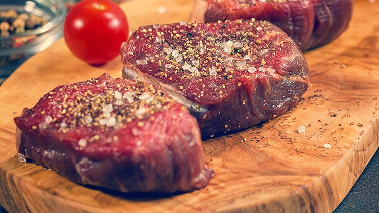 Drei gewürzte Rindersteaks mit Salz und Pfeffer liegen auf einem hölzernen Schneidebrett neben einer roten Kirschtomate.