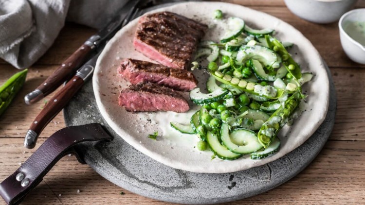 Ein Teller mit mittig gebratenem Steak in Scheiben, Gurkensalat mit Erbsen und Dressing, daneben Messer und kleine Schalen.