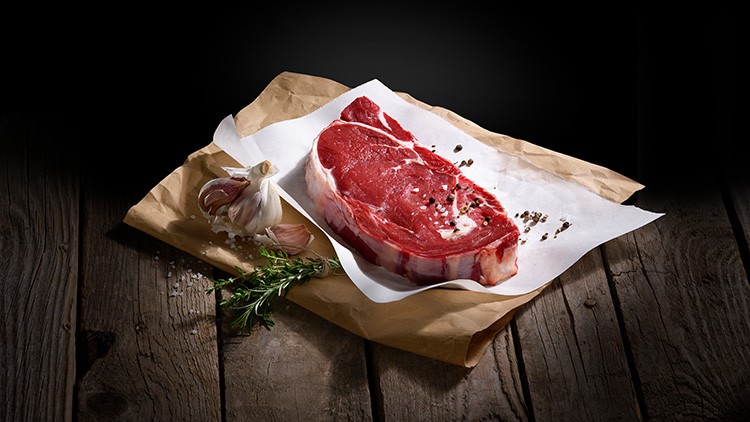 Ein rohes Rindersteak auf weißem Papier mit Knoblauch, Rosmarin, grobem Salz und Pfeffer, auf Holzuntergrund arrangiert.