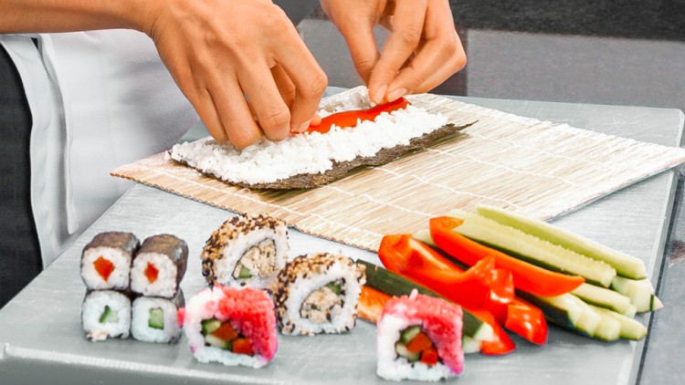 Hände rollen Sushi mit Reis und Paprika auf einem Nori-Blatt, daneben fertige Sushirollen, Gurken- und Paprikastreifen.