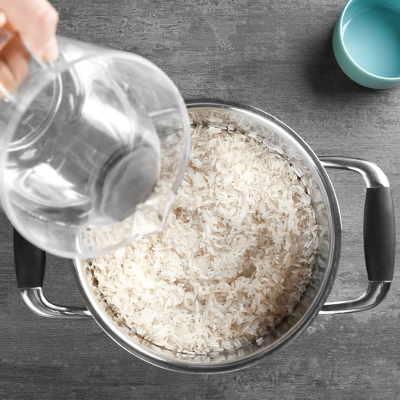 Eine Hand gießt Wasser aus einem transparenten Messbecher in einen Topf mit ungekochtem Reis auf grauer Arbeitsfläche.