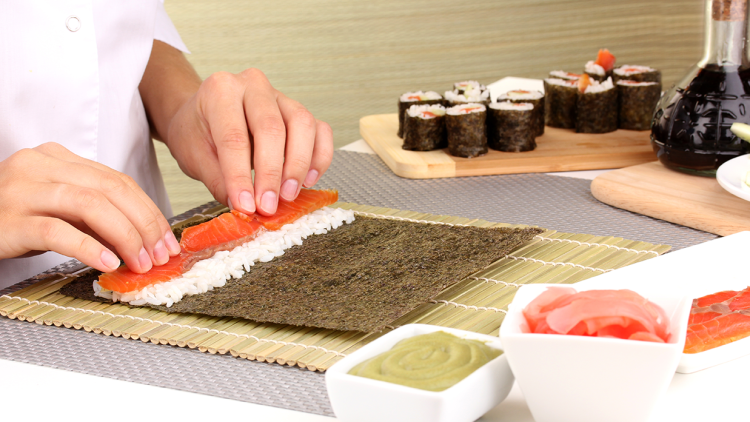 Hände rollen eine Sushirolle mit Lachs, Reis und Algen auf einer Bambusmatte, daneben Wasabi, Ingwer und fertiges Sushi.