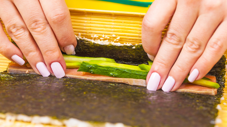 Hände rollen mit einer Bambusmatte ein Nori-Blatt mit Gurken, Lachs und Sesam zu einer Sushi-Rolle.