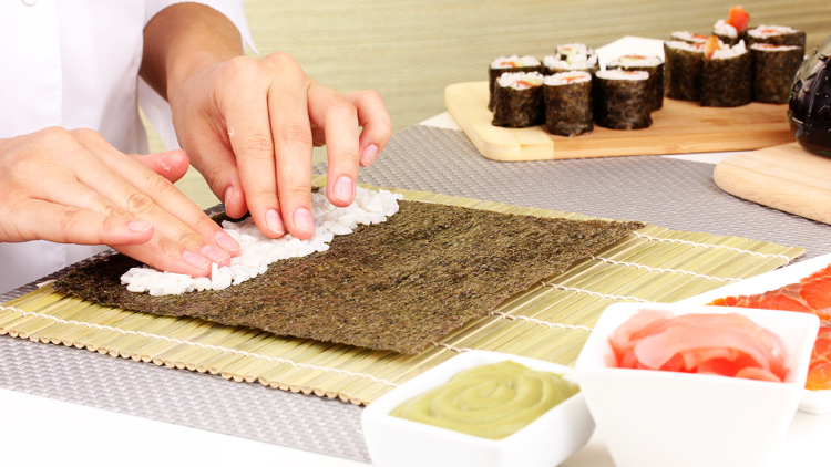 Hände verteilen Reis auf einem Noriblatt auf einer Bambusmatte, daneben Sushi, Ingwer, Wasabi und Lachs in Schalen.
