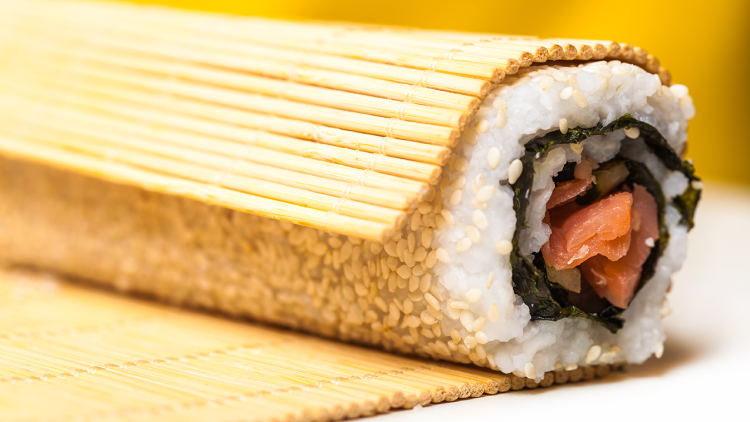Nahaufnahme einer halbgerollten Sushirolle mit Reis, Sesam, Algenblatt und Lachs auf einer Bambusmatte.
