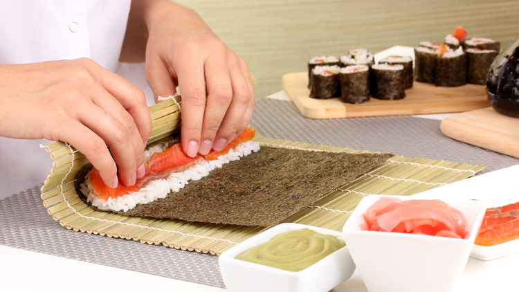 Hände rollen auf einer Bambusmatte ein Sushi aus Reis, Norialge und Lachs, im Vordergrund Wasabi und eingelegter Ingwer.