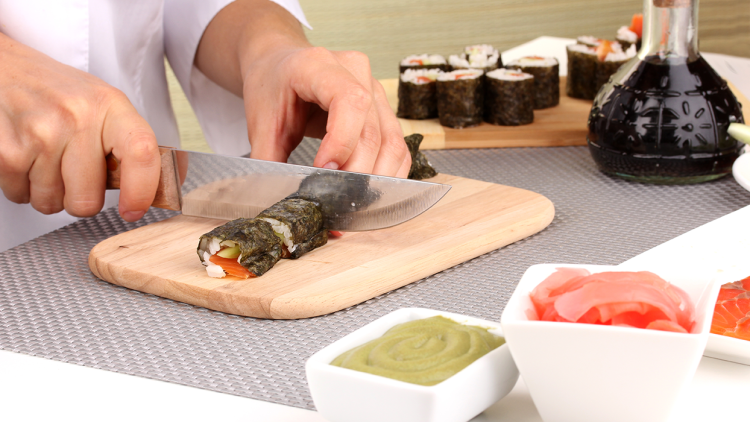 Hände schneiden eine Sushirolle mit einem Messer auf einem Holzbrett, daneben Wasabi, Ingwer und Sojasauce in Schalen.
