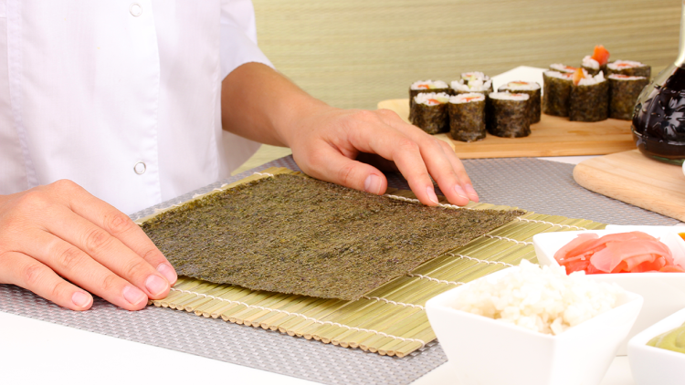 Hände legen ein Nori-Blatt auf eine Bambusmatte, im Hintergrund Sushiröllchen, Reis, Ingwer und Wasabi in Schälchen.
