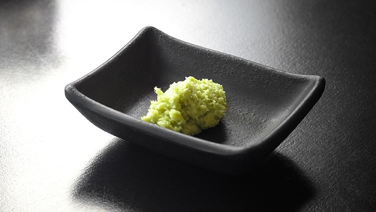 Eine Portion hellgrüner Wasabi liegt in einer kleinen schwarzen Schale auf einer glänzenden dunklen Tischoberfläche.