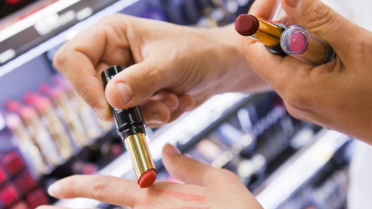 Zwei Hände testen rote Lippenstifte auf dem Handrücken, im Hintergrund stehen weitere Lippenstifte in Regalen.