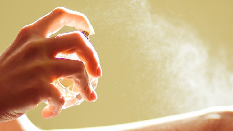 Eine Hand sprüht Parfüm aus einem kleinen Glasflakon, der feine Nebel ist vor einem gelblichen Hintergrund sichtbar.