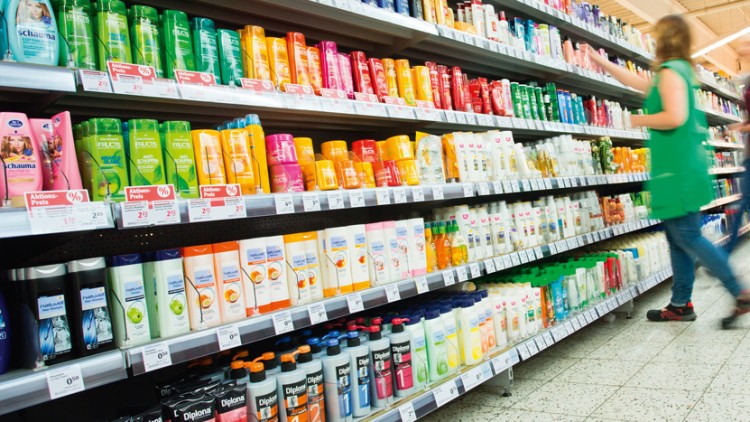 Mehrere Regalreihen mit bunten Shampooflaschen und Pflegeprodukten, eine Person in grüner Kleidung geht vorbei.