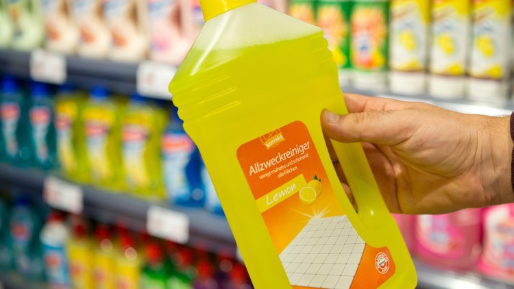 Eine Hand hält eine gelbe Flasche Allzweckreiniger mit der Aufschrift „Lemon“ vor einem Regal mit weiteren Reinigungsmitteln.