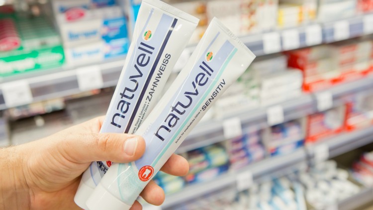 Hand hält zwei Zahnpastatuben der Marke natuvell mit den Varianten ZAHNWEISS und SENSITIV vor einem Regal mit Zahncremes.