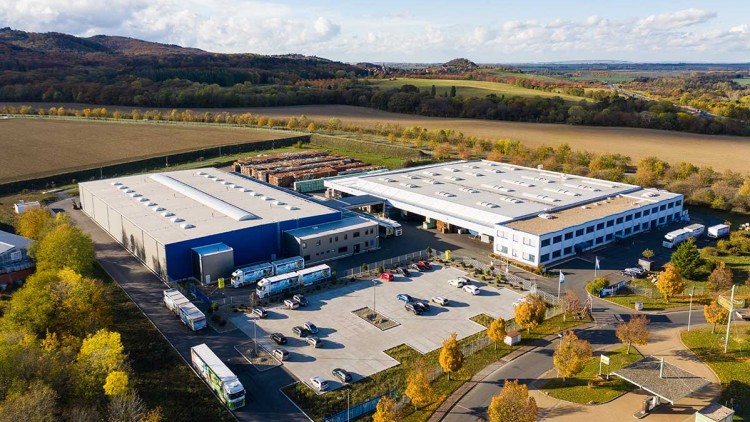 Industriegebäude mit blauen und weißen Fassaden, geparkten Lkws und Pkw vor einem Hintergrund aus Feldern und Wäldern.