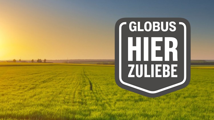 Sonnenaufgang über einer grünen Wiese, rechts schwarzes Schild mit weißer Aufschrift „GLOBUS HIER ZULIEBE“.